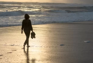 Uma mulher andando sozinha descalça na praia, simbolizando a alma que teme recomeçar.