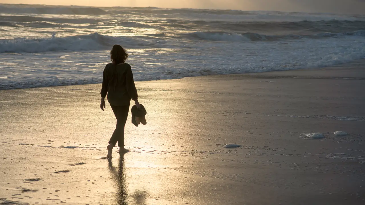 Uma mulher andando sozinha descalça na praia, simbolizando a alma que teme recomeçar.