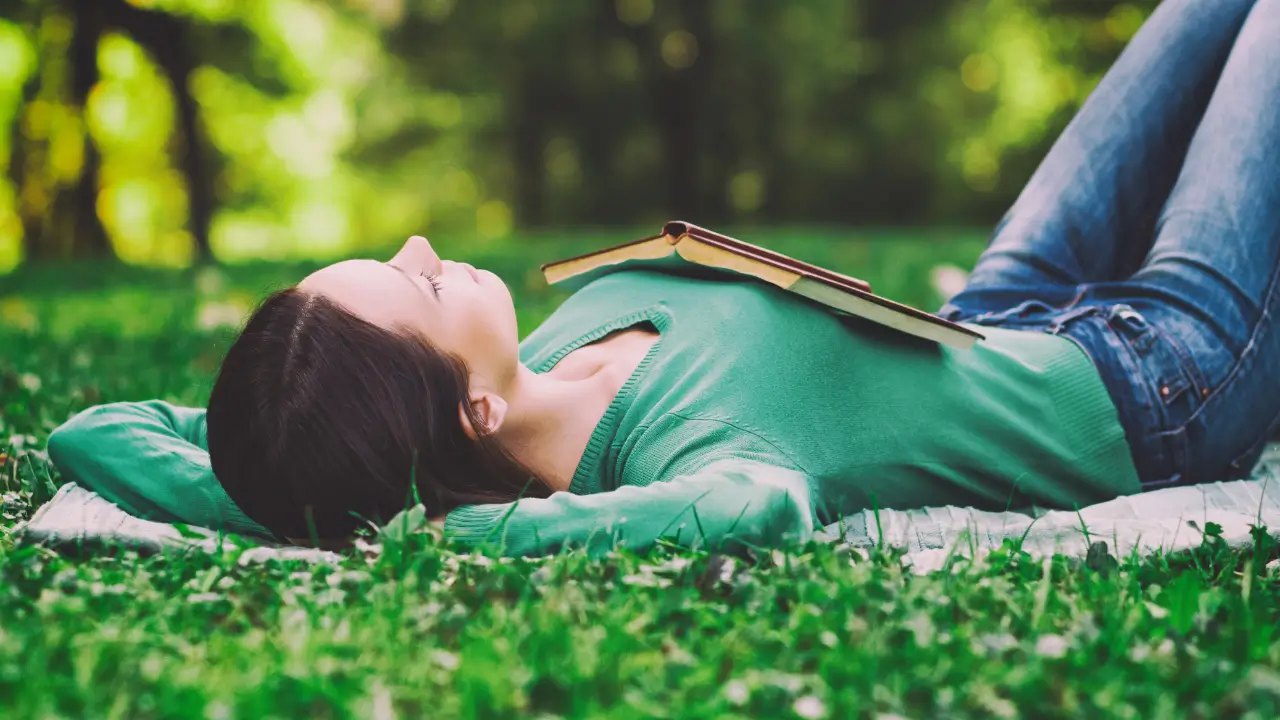 Uma mulher deitada tranquilamente na grma com um livro sobre o colo para simbolizar que descansar é um ato de fé.
